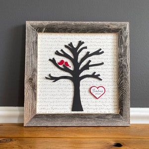 May include: A framed black silhouette of a tree with two red birds perched on a branch. A red heart with the text "Simmons 2024" is attached to the tree. The background is a white paper with cursive writing.