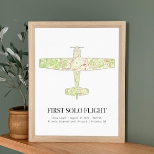 May include: A framed print featuring a green map-style airplane silhouette. The text "FIRST SOLO FLIGHT" is below the image, along with the name Anna Lopez, date, aircraft tail number, and airport location.