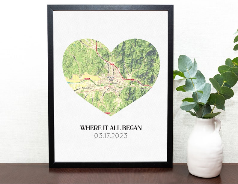 May include: Framed print featuring a heart-shaped map of Grants Pass, Oregon, in shades of green and tan. The text "WHERE IT ALL BEGAN 03.17.2023" is printed below the map in a simple font. The print is in a black frame.