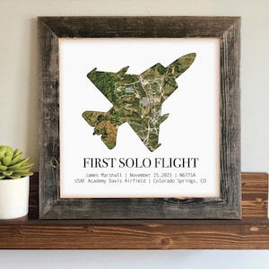 May include: A framed print featuring a detailed aerial view of an aircraft, with the text "FIRST SOLO FLIGHT." The image is set against a white background, framed in rustic wood. Additional text includes a name, date, and location.