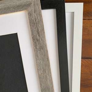 May include: Three picture frames in different colors and styles. The frame on the left is a light gray wood with a white mat. The frame in the middle is black with a white mat. The frame on the right is white with a white mat.