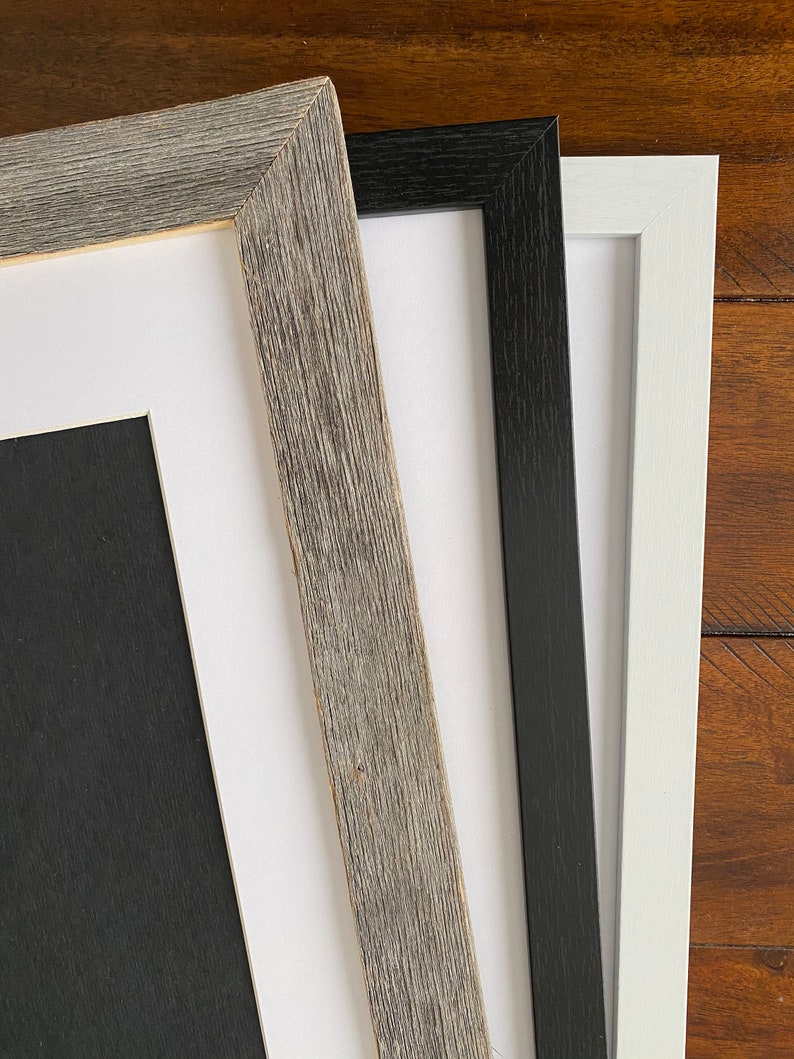 May include: Three picture frames in different colors and styles. The frame on the left is a light gray wood with a white mat. The frame in the middle is black with a white mat. The frame on the right is white with a white mat.