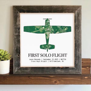 May include: A framed print featuring a green airplane silhouette over a white background. The text "FIRST SOLO FLIGHT" is displayed below the image, along with details about the flight. The frame is made of rustic, weathered wood.