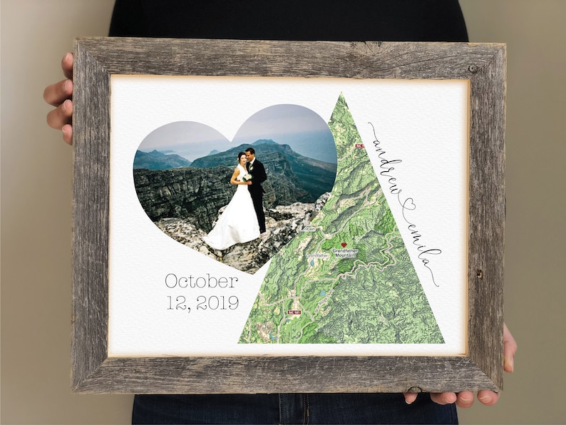 Op de afbeelding: Een ingelijst afdruk met een hartvormige foto van een stel op een bergtop, een kaart van het gebied en de tekst "October 12, 2019" en "andrew & emilia".