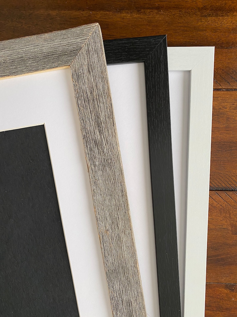 Op de afbeelding: Drie fotolijsten in verschillende kleuren en stijlen. De lijst links is van licht grijs hout met een rustieke afwerking. De middelste lijst is zwart met een gladde afwerking. De lijst rechts is wit met een gladde afwerking.