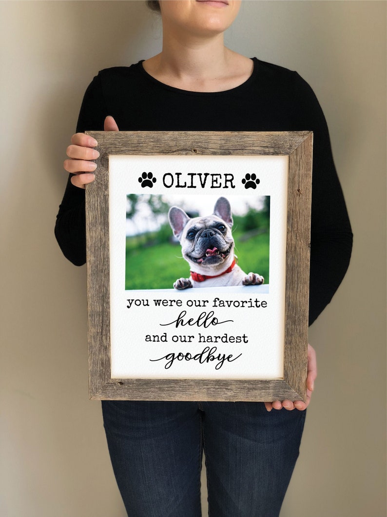 May include: A framed print with a photo of a dog and the text "Oliver" with paw prints on either side. The text below the photo reads "You were our favorite hello and our hardest goodbye".