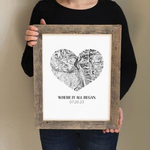 May include: Framed black and white heart-shaped map print with the text "WHERE IT ALL BEGAN 07.20.23". The print is in a rustic, light brown wooden frame. The map details are visible within the heart shape.