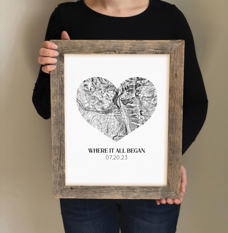 May include: Framed black and white heart-shaped map print with the text "WHERE IT ALL BEGAN 07.20.23". The print is in a rustic, light brown wooden frame. The map details are visible within the heart shape.