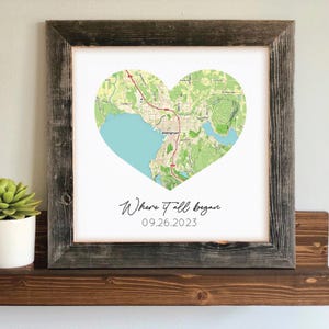 May include: A framed print featuring a heart-shaped map with a light blue water area and green land details. The text "Where it all began" and the date 09.26.2023 are below the map. The frame is made of weathered wood.