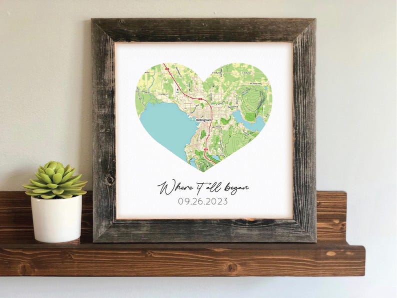 May include: A framed print featuring a heart-shaped map with a light blue water area and green land details. The text "Where it all began" and the date 09.26.2023 are below the map. The frame is made of weathered wood.