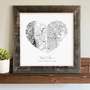 May include: A framed print featuring a heart-shaped map design. The map is split into two sections, each with a different geographical area. The print includes the names "Tim & Lea" and the phrase "LOVE FINDS A WAY" in elegant script. The frame is made of weathered wood.
