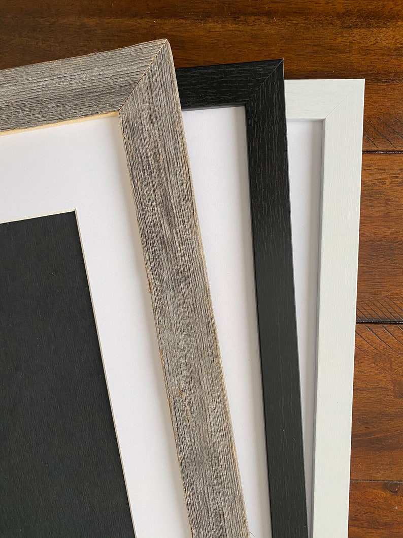 May include: Close-up of three picture frames in different colors: weathered gray, black, and white. The frames have a simple, modern design with a matte finish. The gray frame has a textured, rustic appearance, while the black and white frames are smooth.