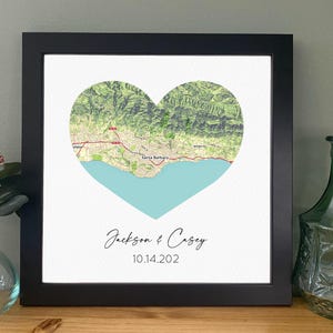 May include: Framed print featuring a heart-shaped map with a coastal landscape and the names "Jackson & Casey" and the date "10.14.202" in elegant script. The map is framed in a black square frame.