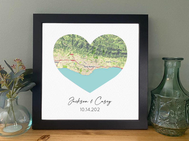 May include: Framed print featuring a heart-shaped map with a coastal landscape and the names "Jackson & Casey" and the date "10.14.202" in elegant script. The map is framed in a black square frame.