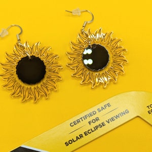 May include: A pair of gold-coloured earrings shaped like suns with mirrored centres. The earrings are on a yellow background with a sign that reads "Certified Safe for Solar Eclipse Viewing Total Eclipse Event Apr. 8 2024".