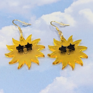 Puede incluir: Un par de pendientes de sol amarillos con gafas de sol negras en forma de estrella. Los pendientes están hechos de acrílico y tienen un gancho dorado.