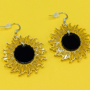 May include: A pair of gold sunflower earrings with black centres. The earrings are made of acrylic and have a simple design.