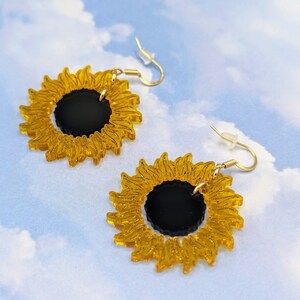 May include: A pair of gold sunflower earrings with black centres. The earrings are made of acrylic and have a delicate, whimsical design.