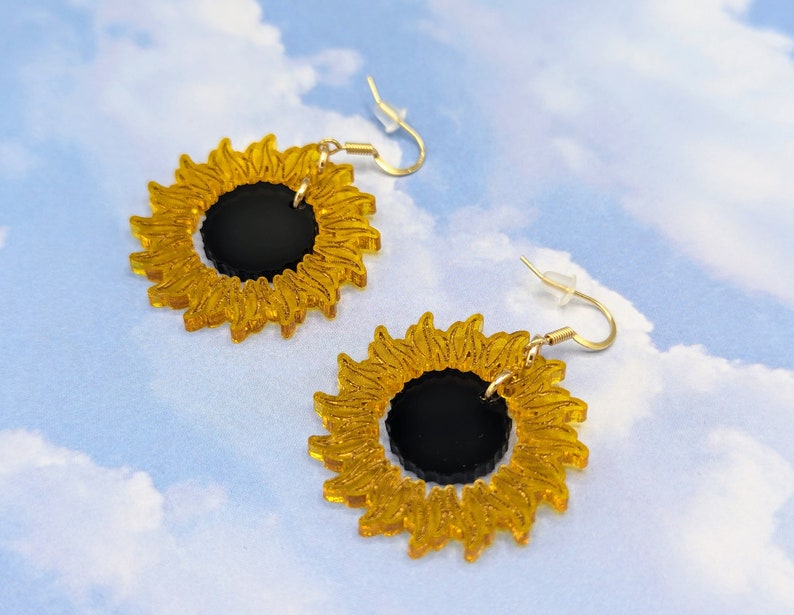 May include: A pair of gold sunflower earrings with black centres. The earrings are made of acrylic and have a delicate, whimsical design.
