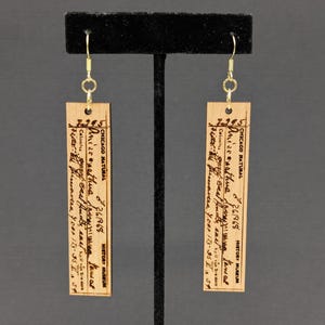 Puede incluir: Un par de pendientes rectangulares de madera con ganchos dorados. Cada pendiente presenta un diseño impreso con el texto "CHICAGO NATURAL HISTORY MUSEUM" y texto escrito a mano. Los pendientes se exhiben en un soporte negro.