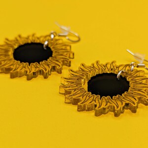 May include: A pair of gold sunflower earrings with black centres. The earrings are made of acrylic and have a delicate, intricate design.