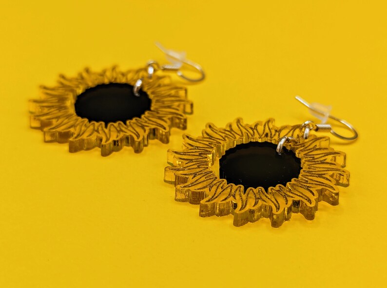 May include: A pair of gold sunflower earrings with black centres. The earrings are made of acrylic and have a delicate, intricate design.