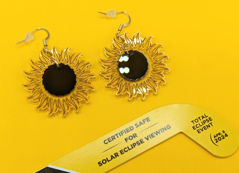 May include: A pair of gold-coloured earrings shaped like suns with mirrored centres. The earrings are on a yellow background with a sign that reads "Certified Safe for Solar Eclipse Viewing Total Eclipse Event Apr. 8 2024".