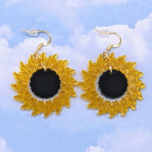 May include: A pair of gold sunflower earrings with black centres. The earrings are made of acrylic and have a shiny finish.