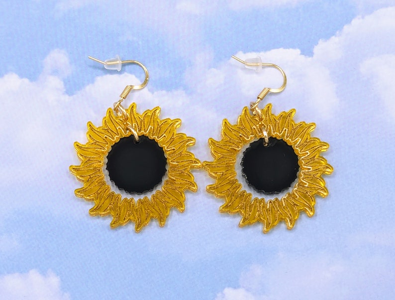 May include: A pair of gold sunflower earrings with black centres. The earrings are made of acrylic and have a shiny finish.