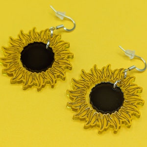 May include: A pair of sunflower earrings with black centres and yellow petals. The earrings are made of acrylic and have silver hooks.