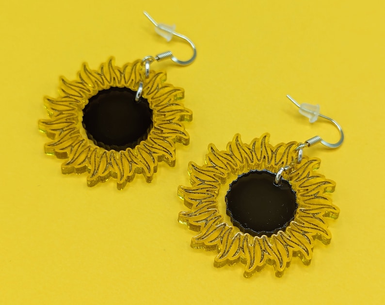 May include: A pair of sunflower earrings with black centres and yellow petals. The earrings are made of acrylic and have silver hooks.