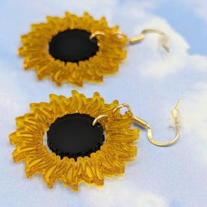 May include: A pair of gold sunflower earrings with black centres. The earrings are made of acrylic and have a delicate, whimsical design.