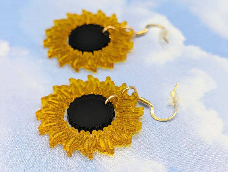 May include: A pair of gold sunflower earrings with black centres. The earrings are made of acrylic and have a delicate, whimsical design.