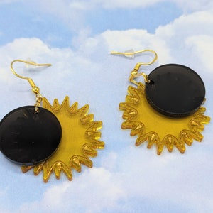 Puede incluir: Un par de pendientes dorados con un círculo negro y un diseño de sol amarillo.