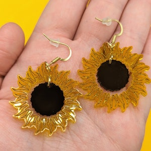 May include: A pair of gold sunflower earrings with black centres. The earrings are dangle style with a hook fastening.