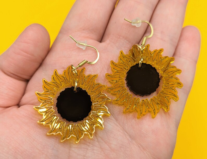 May include: A pair of gold sunflower earrings with black centres. The earrings are dangle style with a hook fastening.