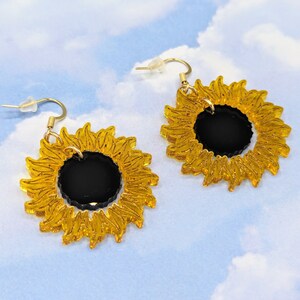 May include: A pair of gold sunflower earrings with black centres. The earrings are made of acrylic and have a simple, modern design.