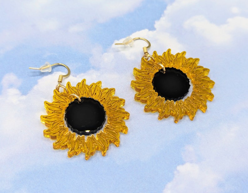 May include: A pair of gold sunflower earrings with black centres. The earrings are made of acrylic and have a simple, modern design.