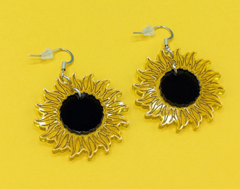 May include: A pair of gold sunflower earrings with black centres. The earrings are made of acrylic and have a simple design.