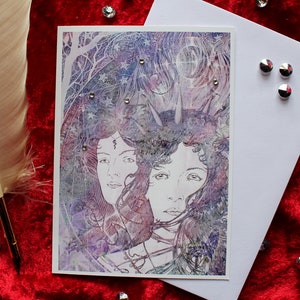 May include: A greeting card with an illustration of two women with long hair, decorated with floral and celestial designs. The artwork uses purple, blue, and white tones, and is embellished with small, sparkling gems. A white envelope and a feather quill are also present.