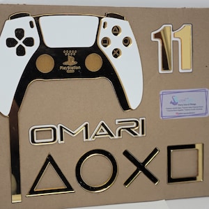 Puede incluir: Un mando de PlayStation blanco y dorado con el logotipo de PlayStation, el nombre "OMARI" en letras doradas, el número "11" en dorado y los símbolos de PlayStation triángulo, círculo, cruz y cuadrado en dorado.