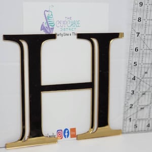 May include: Gold mirrored letter H, approximately 7 inches tall, with a black backing. The letter is on a white surface with a ruler for scale.
