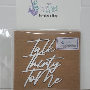 Op de afbeelding: Witte vinyl taarttopper met de woorden "Talk Thirty To Me" in een cursief schrift. De topper is verpakt op een bruine kartonnen achtergrond met een klein wit label waarop "The Cupcake District Party Line & Things" staat.