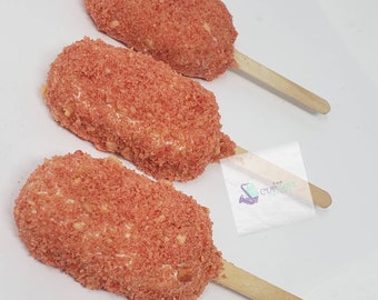 May include: Three strawberry shortcake pops on wooden sticks. The pops are covered in a red and white crumb topping.
