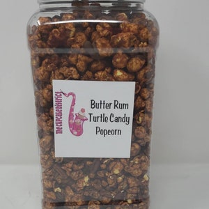 May include: A clear plastic jar filled with brown and tan popcorn. The jar has a black lid and a white label that says "Butter Rum Turtle Candy Popcorn" with a pink and purple saxophone graphic.