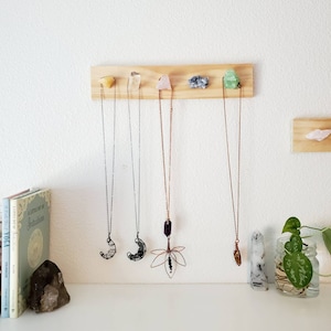 May include: A wooden display board with five crystal necklaces hanging from it. The necklaces feature various gemstones, including a moon-shaped pendant, a lotus flower pendant, and other crystal pendants. The board is mounted on a white wall.