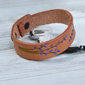 May include: A tan leather bracelet with a floral design. The bracelet features hand-painted purple flowers and green stems. The bracelet has a snap closure and the text "Lightning Bug Creations LLC".