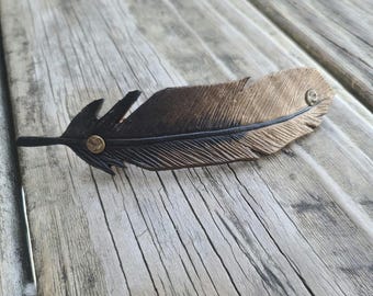 May include: A handcrafted leather feather hair clip. The feather is primarily brown with a black stem and tip. A thin black line runs down the center of the feather. The feather is secured with two small brass rivets. The clip is resting on a weathered wooden surface.