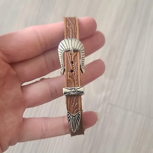 May include: A brown leather bracelet with a silver-colored buckle and accents. The leather has a textured pattern. The buckle is a decorative design. The bracelet is a narrow width.