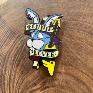 May include: A colorful enamel pin featuring a cartoon rabbit with blue fur, holding a guitar. The pin has the words "BONNIE LOVER" on banners. The guitar is black and yellow. The rabbit has a purple hat.